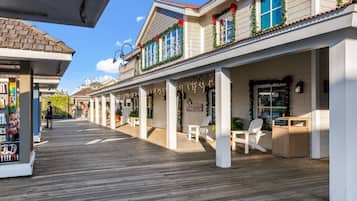 Una terraza de madera cubierta con sillas y mesas blancas, una tienda a la izquierda y un edificio con ventanas adornadas con plantas y listones rojos.