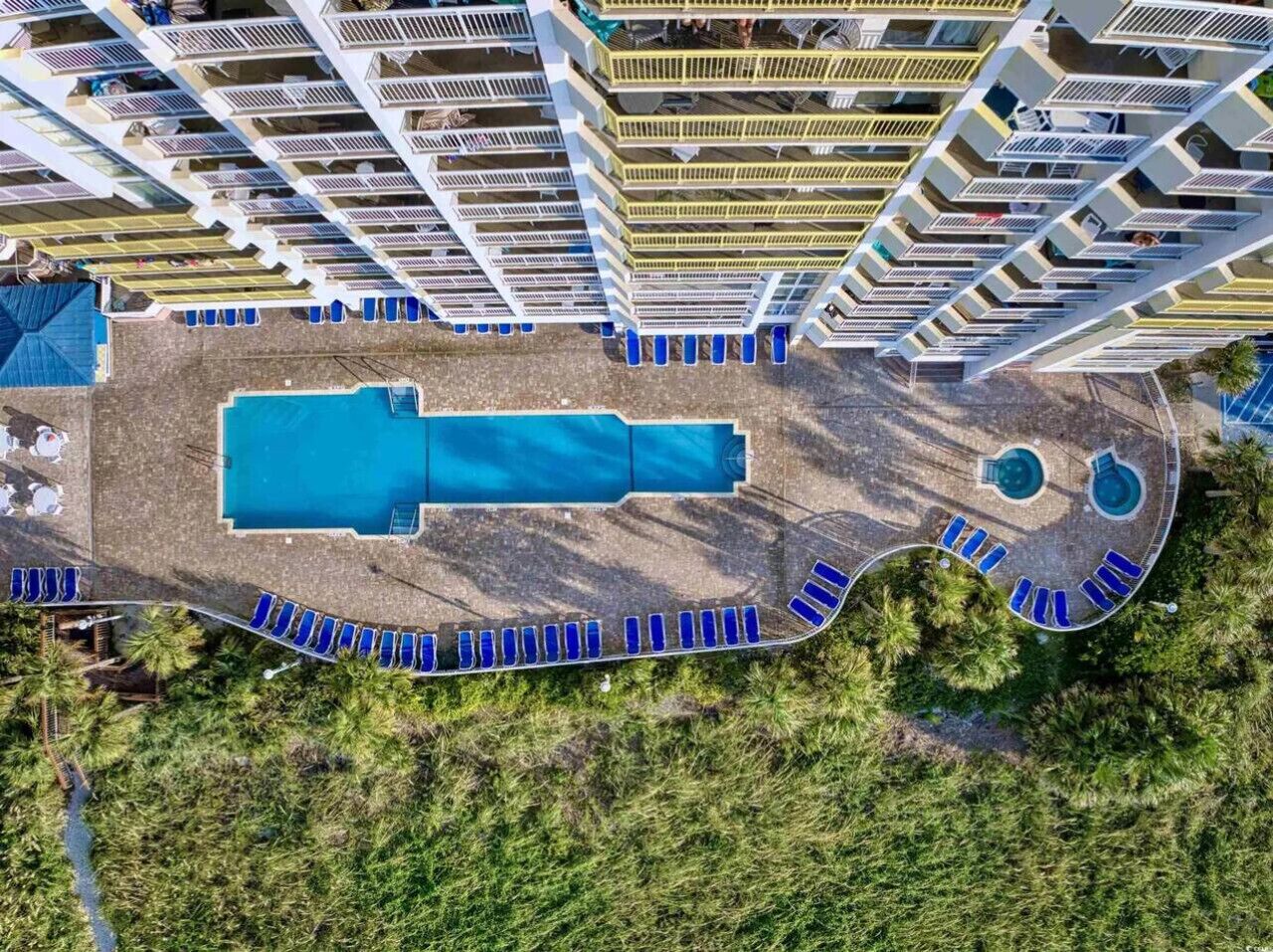 Vue aérienne d’un hôtel doté d’une grande piscine centrale, de nombreux balcons et d’un espace piscine aménagé avec des chaises longues.