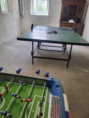 A ping pong table and a foosball table in a room with a window and a wooden cabinet.