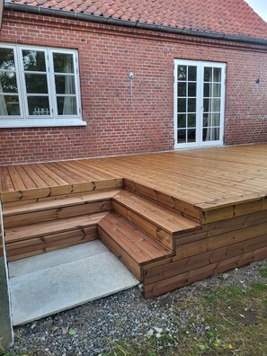 A wooden deck with steps leading to a brick building with two white-framed windows.