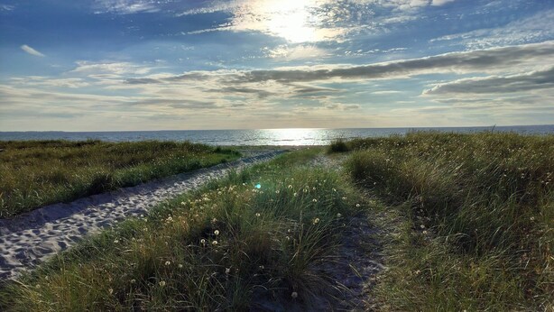 Ein Küstenpfad mit Sand und Gras, der zum Meer unter einem teils bewölkten Himmel führt.