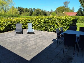 An outdoor patio area with lounge chairs and a table, surrounded by well-maintained hedges and trees. - Lovely holiday home near sandy beach (Vig)