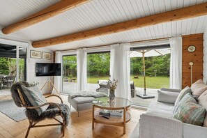 A cozy living room with a wooden ceiling, a TV, a sofa, a chair, and a view of a garden. - Danish summerhouse with Danish Holiday Atmosphere (Væggerløse)