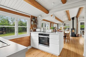 A modern kitchen with wooden beams, a central island, a built-in oven, and a large window overlooking a garden. - Danish summerhouse with Danish Holiday Atmosphere (Væggerløse)