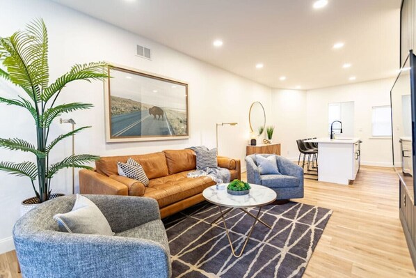 A modern living room with a sofa, armchairs, a coffee table, a large plant, a painting, and a kitchen area with a sink and counter.