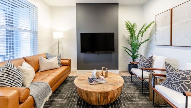 A modern living room with a leather sofa, a round coffee table, a TV mounted on a dark wall, and a potted plant. - 2BR Home | Near T | Free Street Parking (Pittsburgh)