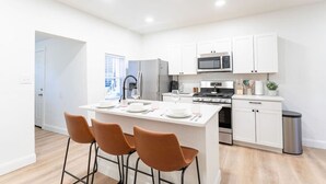 A modern kitchen with white cabinets, stainless steel appliances, a central island with bar stools, and a view of the city through the window. - 2BR Home | Near T | Free Street Parking (Pittsburgh)