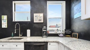 Une cuisine dotée d’un plan de travail en granit, de meubles blancs, d’un lave-vaisselle, d’une machine à café et d’une fenêtre offrant une vue sur une maison et l’eau.