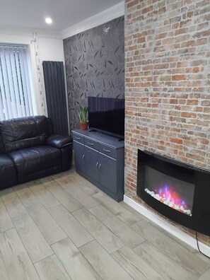 Ein Wohnzimmer mit Kamin aus Ziegelsteinen, einer Ledersofa, einem Fernseher auf einem Schrank und einer Wand mit Muster.