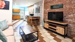 A living room with a TV, a sofa, a coffee table, a kitchen in the background, and a brick wall. - 1 Block from Butler St ★ Patio ★ Dog Friendly! (Pittsburgh)