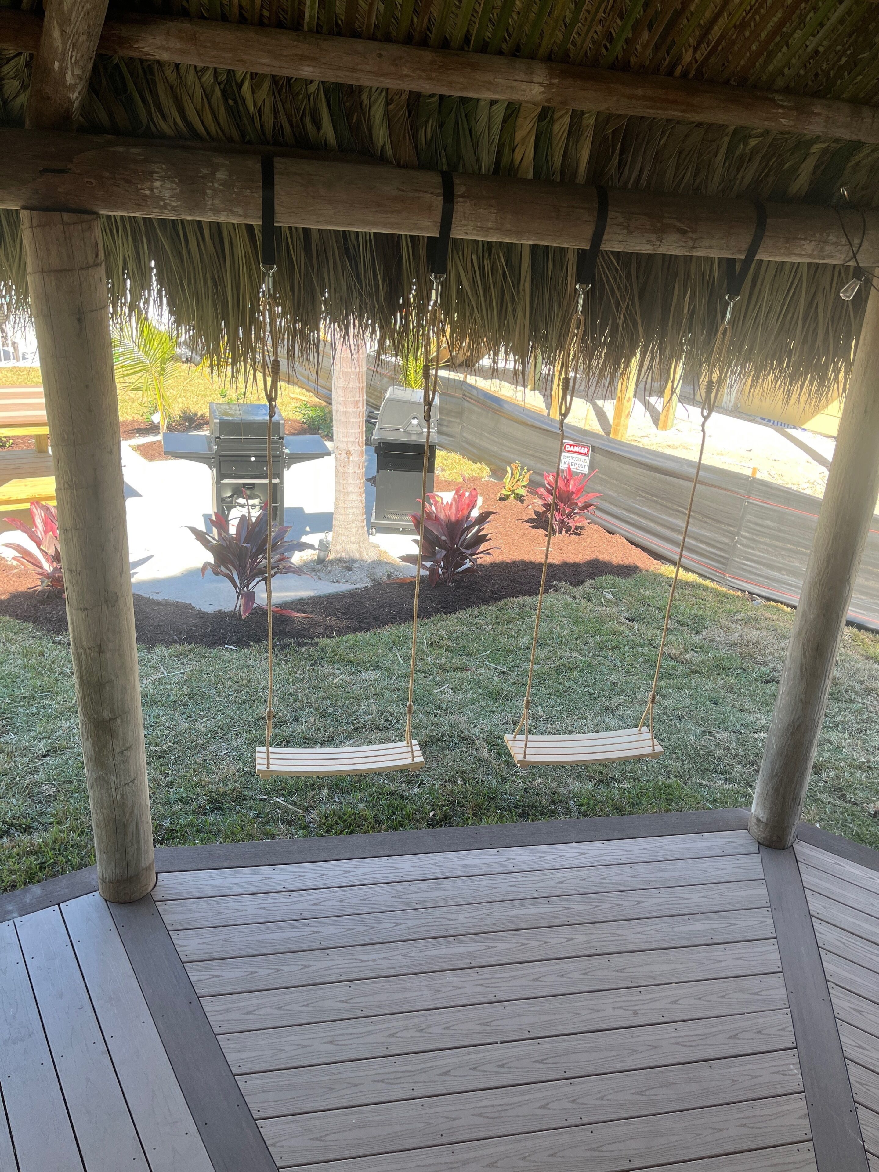A wooden deck with a thatched roof, two swings, a barbecue grill, and a garden with red flowers.