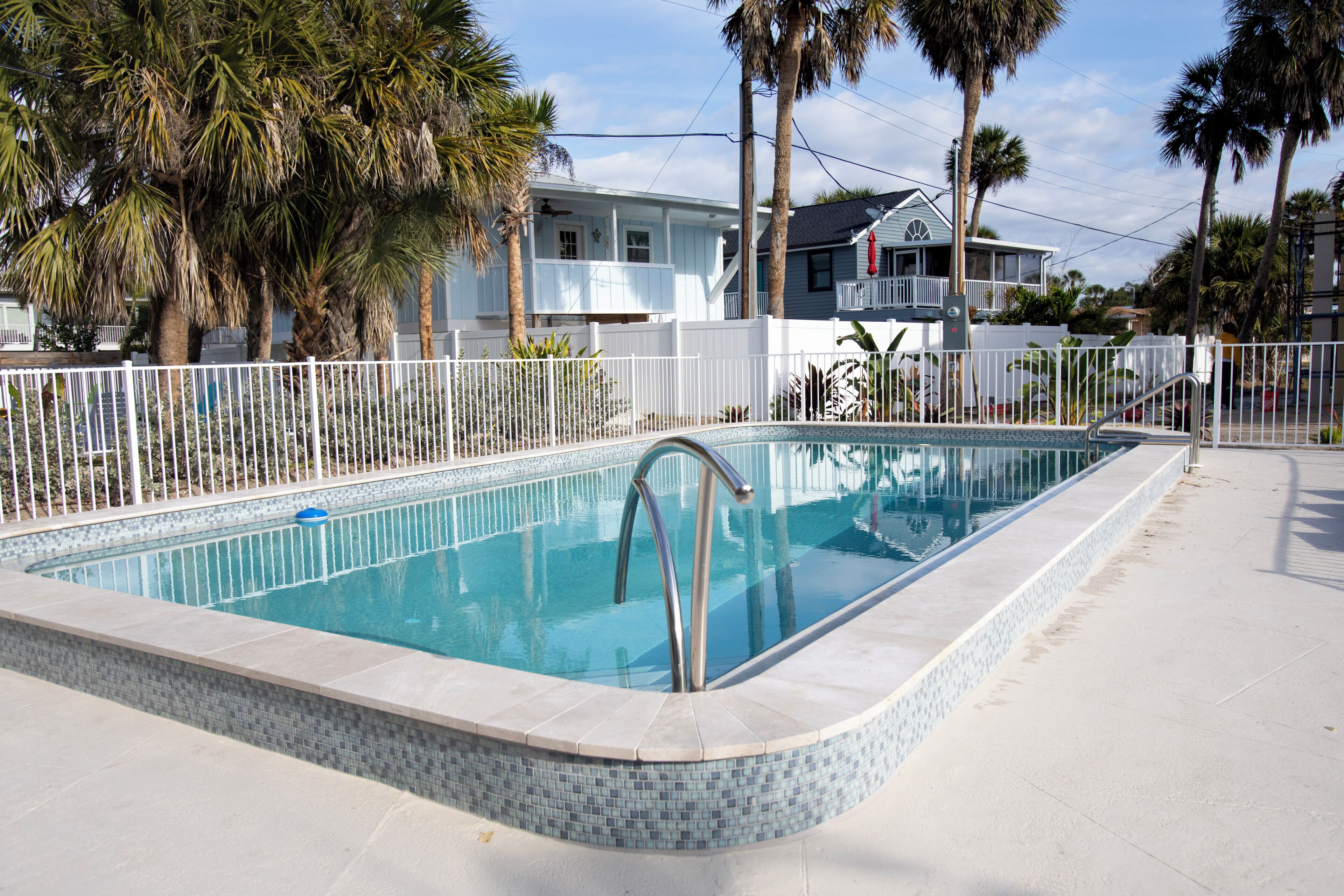 A rectangular swimming pool with a tiled edge, surrounded by a white fence, palm trees, and houses in the background.