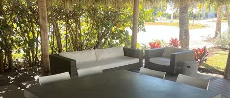 An outdoor seating area with a large table and white chairs, surrounded by a thatched roof structure and lush greenery.