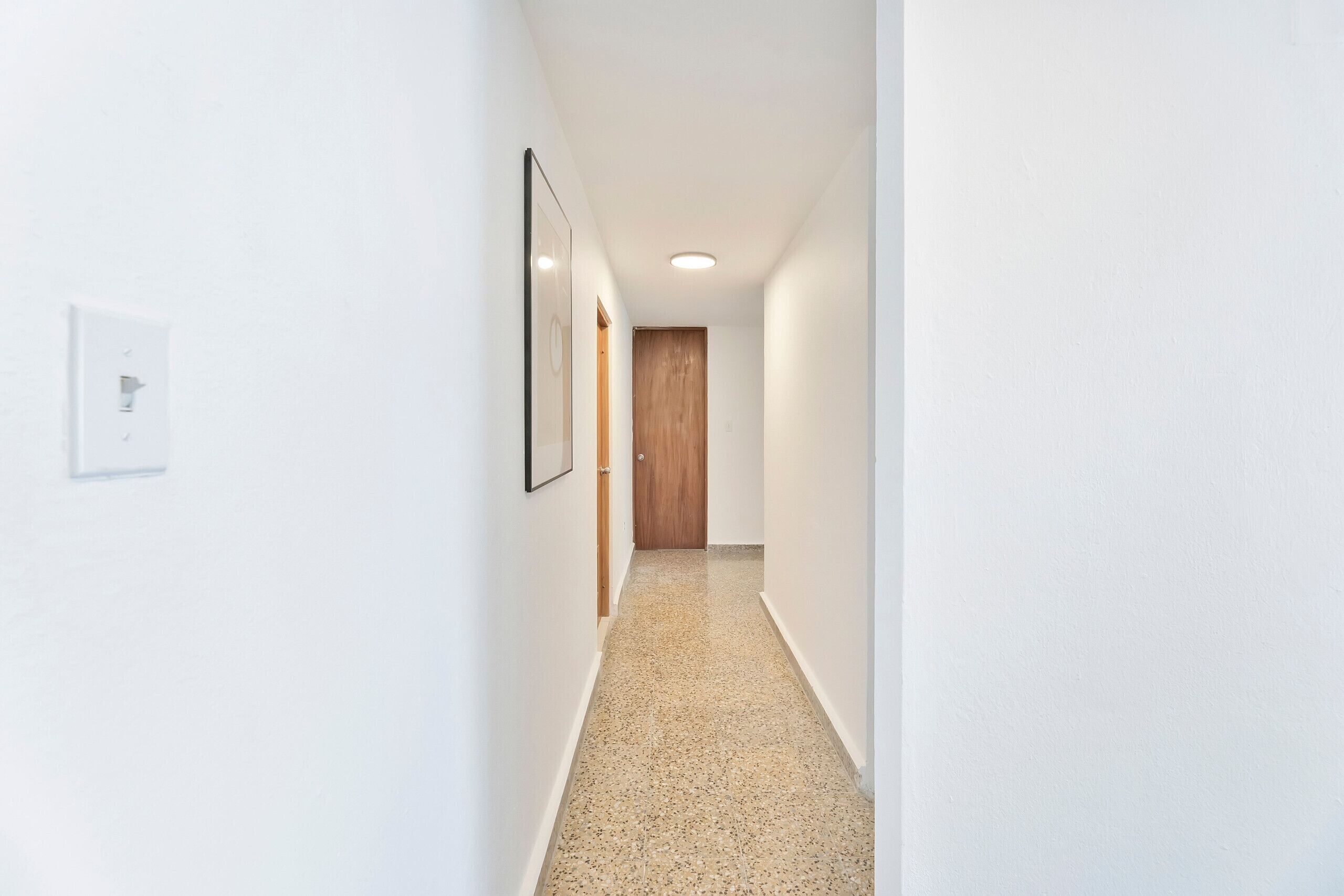 Un couloir étroit aux murs blancs, une porte en bois au fond et un interrupteur sur le côté gauche.