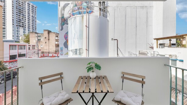 A balcony with a wooden table and two chairs, a potted plant, and a view of a cityscape with buildings and a large cylindrical structure. - Calle Cerra Getaway 3BR w/ Balcony (San Juan)
