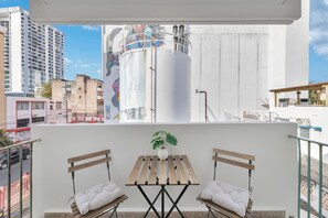 A balcony with a wooden table and two chairs, a potted plant, and a view of a cityscape with buildings and a large cylindrical structure. - Calle Cerra Getaway 3BR w/ Balcony (San Juan)