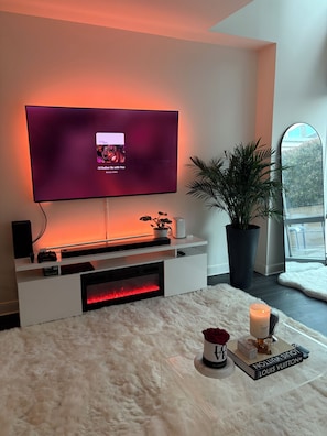 Una sala de estar moderna con un televisor de pantalla plana montado en la pared, un mueble blanco para entretenimiento debajo, un tapete blanco y mullido, una planta en maceta, una vela y un libro Louis Vuitton.