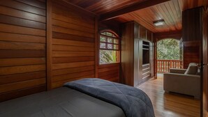 A room with wooden paneling, a bed, a window, and a balcony with a chair. - Casa com Lareira e 3 su tes no Parque das Fontes (Campos do Jordão)