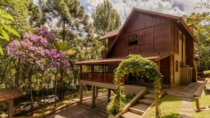 A wooden house with a porch, surrounded by trees and a garden. - Casa com Lareira e 3 su tes no Parque das Fontes (Campos do Jordão)