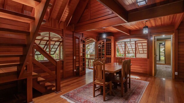 A wooden interior with a staircase, a dining table with chairs, and large windows. - Casa com Lareira e 3 su tes no Parque das Fontes (Campos do Jordão)