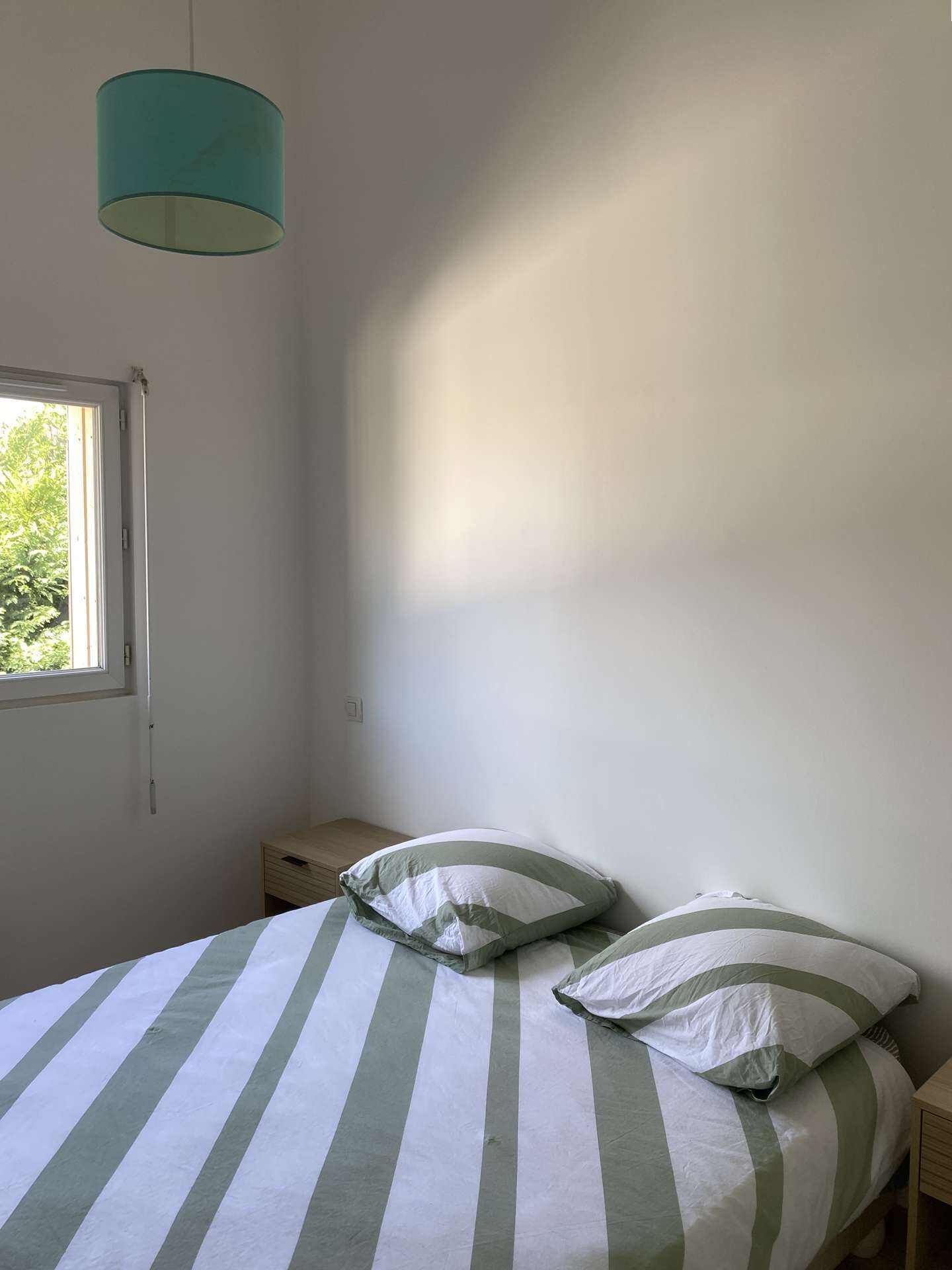 Une chambre à coucher avec un lit, des oreillers, une table de chevet, une fenêtre et un luminaire suspendu.