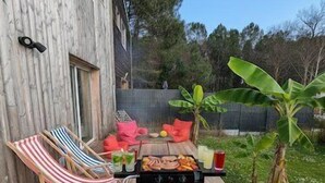 Une terrasse extérieure avec une plateforme en bois, un barbecue, une table garnie de nourriture et de boissons, et un fauteuil de jardin à rayures.