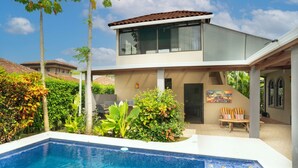 A modern two-story house with a swimming pool, surrounded by lush greenery and palm trees.