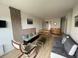 A living room with a sofa, a wooden chair, a dining table with benches, a kitchen area, and a mounted TV. - Seignosse océan, 2 bedroom apartment, walking distance to the beach, ideal for a family (SEIGNOSSE)