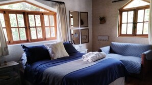 Un dormitorio con una cama, una mesita de noche, una silla y dos ventanas.