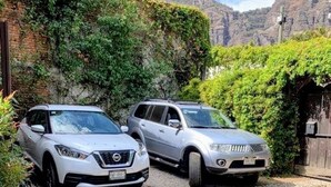 Dos coches estacionados en un camino adoquinado, con vegetación y un fondo rocoso.