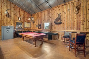 A pool table in a room with wooden walls, a safe, and mounted animal heads.