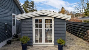 En liten, mörkfärgad byggnad med en vit dörr och vita fönster, två krukväxter och en gång av tegelstenar.