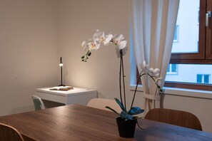 A room with a wooden table, a potted orchid, a desk with a lamp, and a window with curtains.