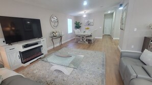 A living room with a flat-screen TV, fireplace, glass coffee table, grey sofa, dining area with chairs, and a hallway. - Beautiful  Vacation Home in The Villages, Florida – Golf Car Included! (Lady Lake)