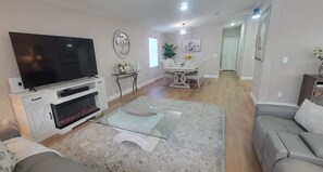 Ein Wohnzimmer mit Flachbildfernseher, Kamin, Glastisch, grauem Sofa, Essbereich mit Stühlen und einem Flur.