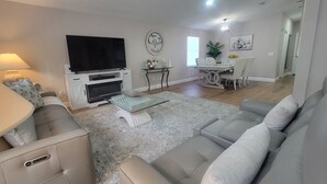 A living room with a sofa, TV, fireplace, dining table, and artwork. - Beautiful  Vacation Home in The Villages, Florida – Golf Car Included! (Lady Lake)