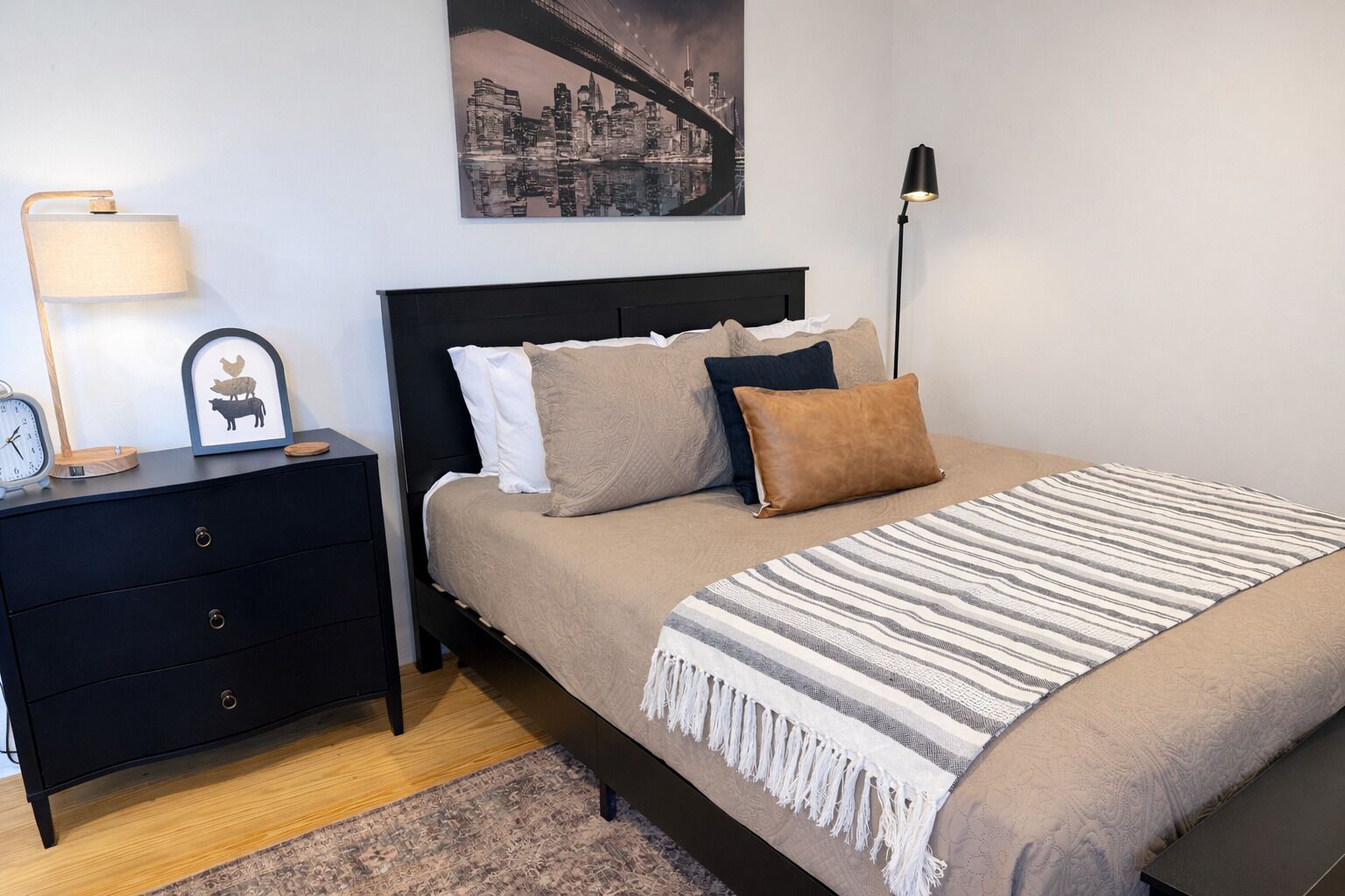 Ein Schlafzimmer mit Bett, Nachttisch, Lampe, gerahmtem Bild und Stadtlandschaftsbild an der Wand.