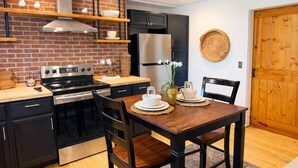 Une cuisine moderne avec un mur en briques, des appareils électroménagers en acier inoxydable, des étagères en bois et un coin repas comprenant une table et des chaises en bois.