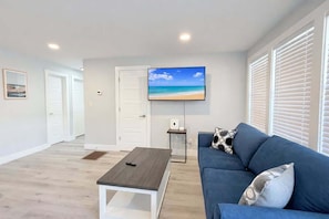 Una sala moderna con un sofá azul, una mesa de centro, un televisor mostrando una playa y puertas blancas.