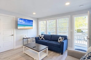 Una sala de estar con un sofá azul, una mesa de centro, un televisor en la pared y ventanas amplias.