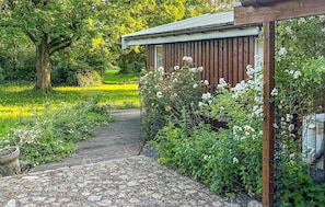 Un sendero en un jardín que lleva a una estructura de madera con techo metálico, rodeada de abundante vegetación y plantas en flor.