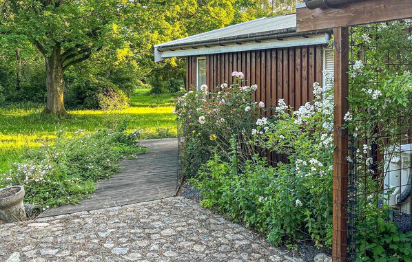 A garden pathway leading to a wooden structure with a metal roof, surrounded by lush greenery and flowering plants.