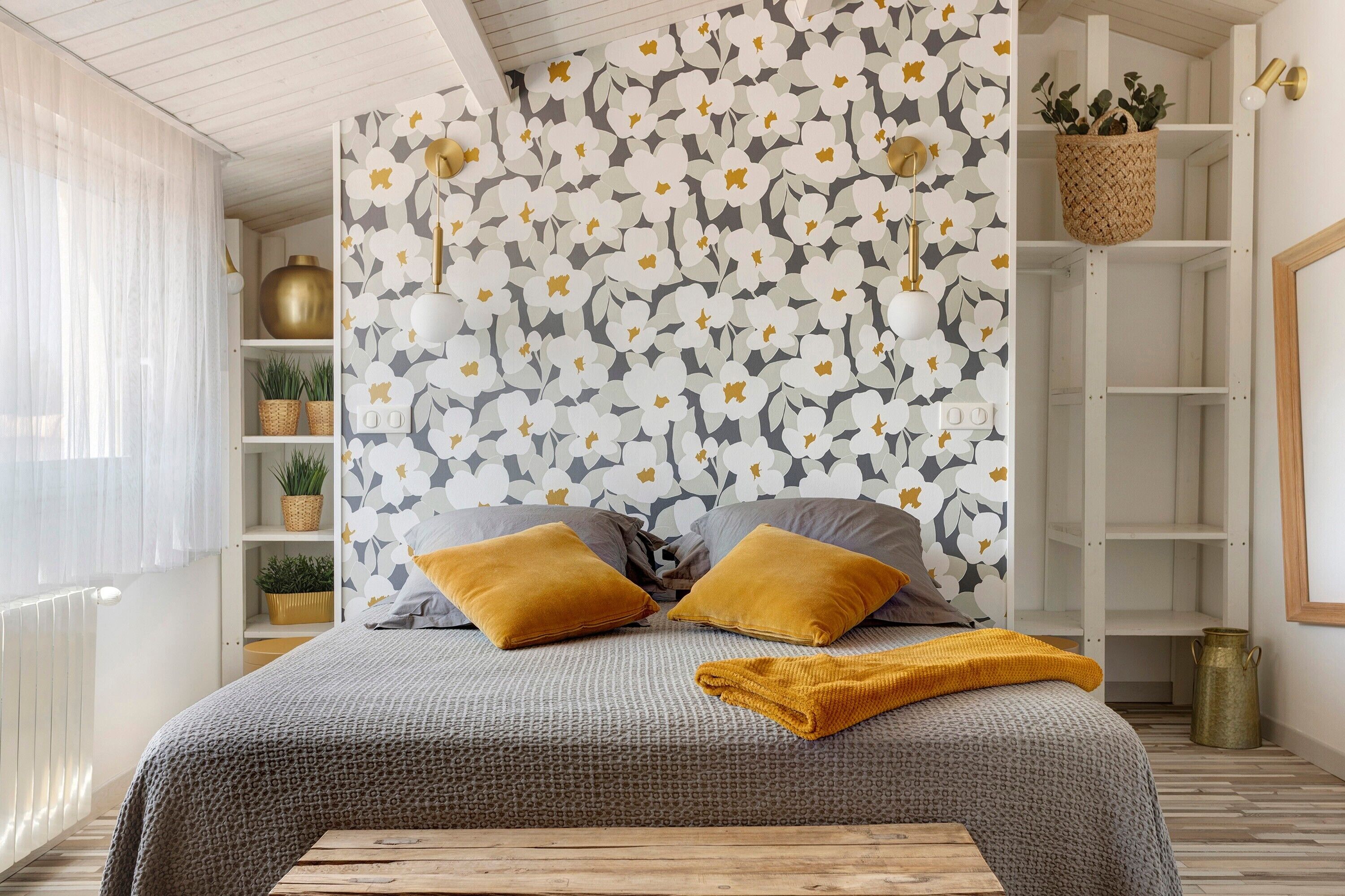 A bedroom with a patterned wallpaper, a bed with two pillows, a bedside table, and a hanging plant.