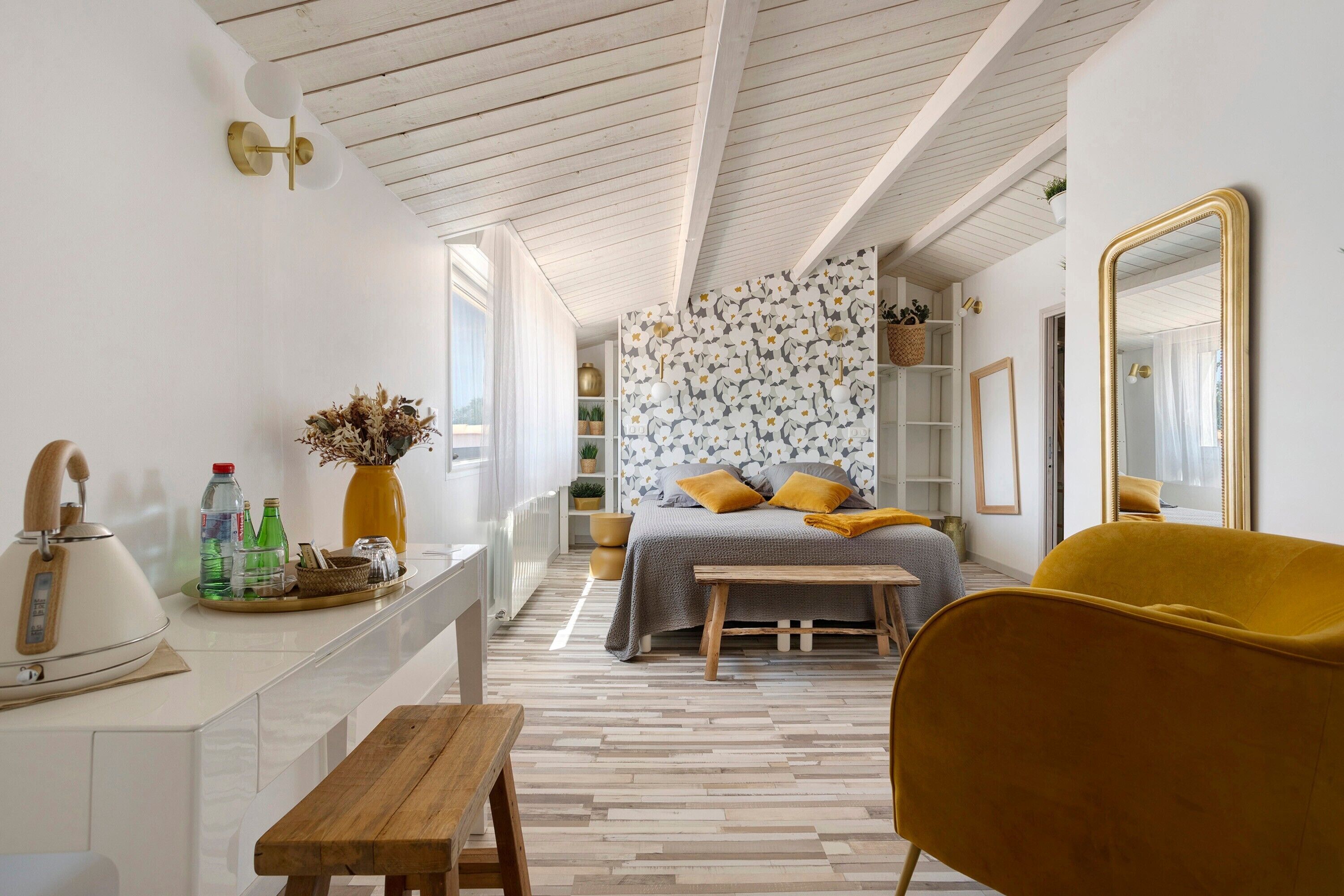 Ein modernes Schlafzimmer mit Holzboden, einem Bett mit gemustertem Kopfteil, einem gelben Sessel und einem Nachttisch mit Wasserkocher und Flaschen.