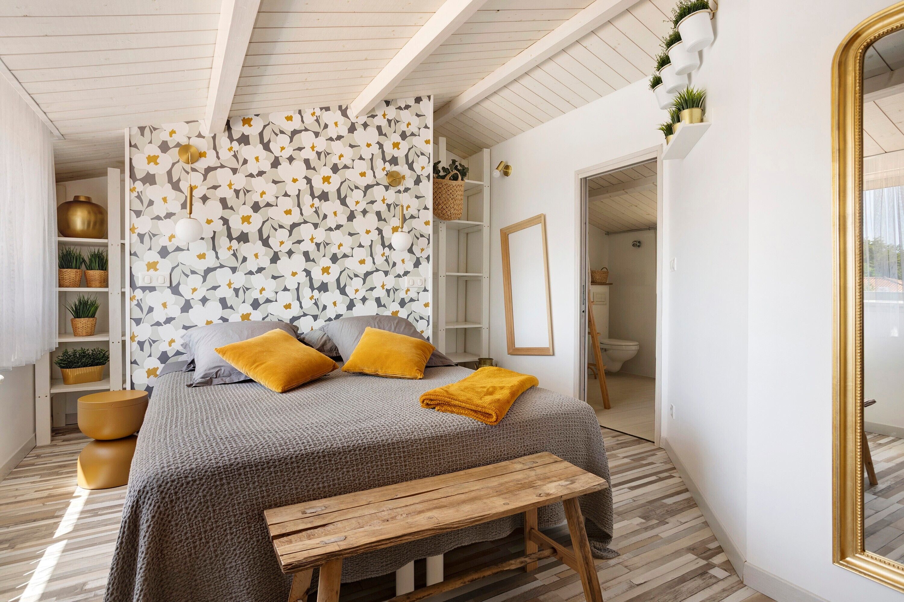 A bedroom with a bed, a bench, a mirror, and a wall with a floral pattern.