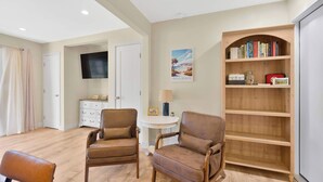 Una habitación con dos sillones de cuero, una estantería de madera, un televisor, una mesita redonda con una lámpara y una puerta que da a otra habitación.