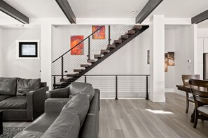 Ein modernes Wohnzimmer mit grauem Ledersofa, einem Esstisch mit Stühlen, einer Treppe mit schwarzer Geländer und abstrakter Wandkunst.