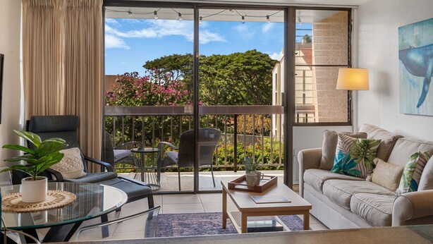 A modern living room with a glass table, a sofa, a chair, and a balcony with a view of trees and buildings. - Kihei Maui Vista Condo | 3 Pools, Walk to Beach, Tennis, Pickleball, AC (Kihei)