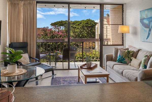 A modern living room with a glass table, a sofa, a chair, and a balcony with a view of trees and buildings. - Kihei Maui Vista Condo | 3 Pools, Walk to Beach, Tennis, Pickleball, AC (Kihei)
