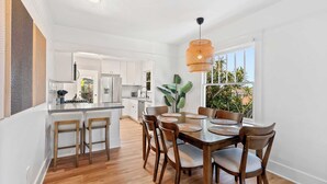 A modern kitchen with white cabinets, a dining area with a wooden table and chairs, and a large window with a view of greenery. - Steps to La Jolla Cove - Private Outdoor Oasis (San Diego)